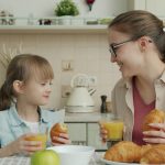Young woman and adorable little girl are eating croissants and drinking juice talking at breakfast at home, modern interior is visible in background.
